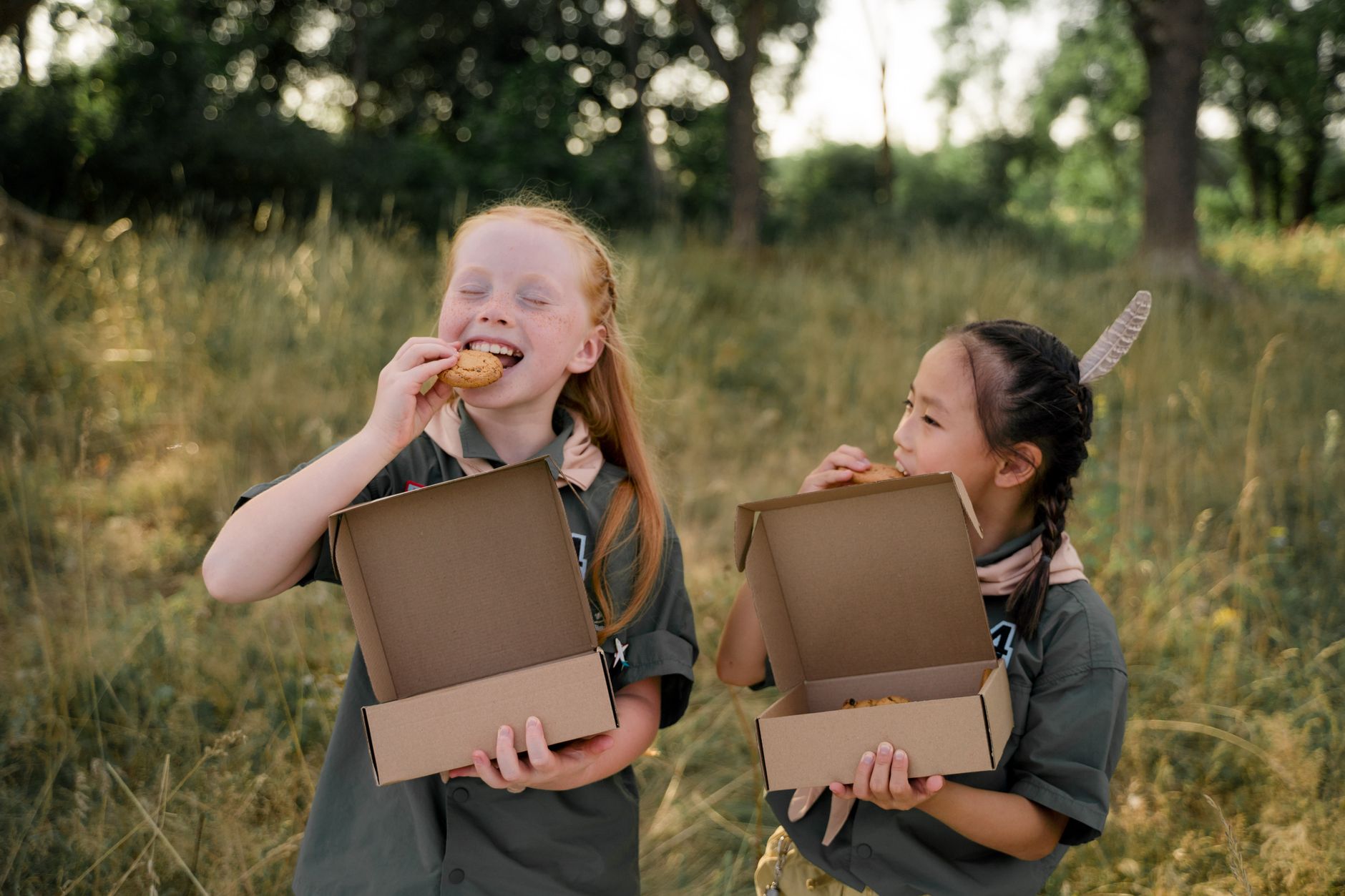 Two girls in scout uniforms enjoying cookies in a grassy outdoor setting.