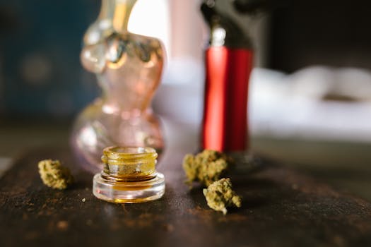 Glass jar with cannabis concentrate, surrounded by dried marijuana buds and smoking accessories on a table.