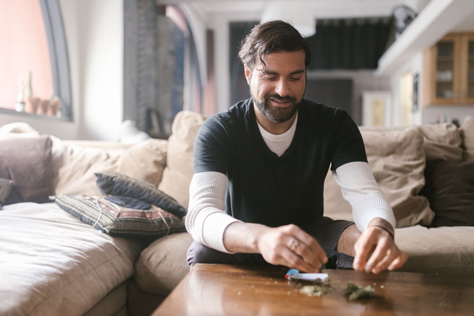 A man sits on a couch rolling a joint in a comfortable living room environment.